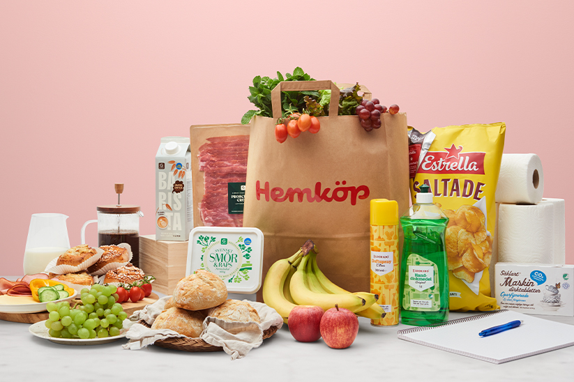 En Hemköp-matkasse med matvaror och chips, bananer, smör, vindruvor, frallor, kaffe, mjölk, äpplen, hushållspapper och mer matvaror står på ett marmorbord framför en rosa bakgrund.
